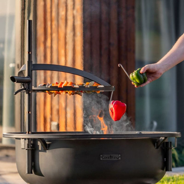 Man lägger paprika på grillgaller ovanför eldfat från Cook King Bandito i kvällssol.