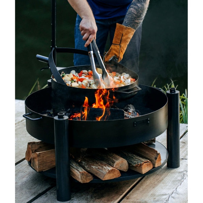 Matlagning i stekpanna på MONTANA X eldfat, med synlig låga och justerbart stöd.