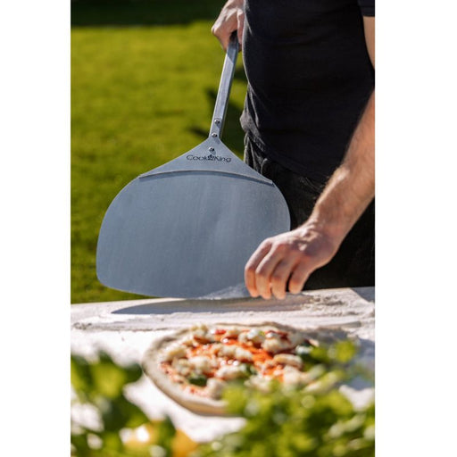 Person håller i en pizzaspade i rostfritt stål utomhus, redo att lyfta en pizza från ett mjölat bord.