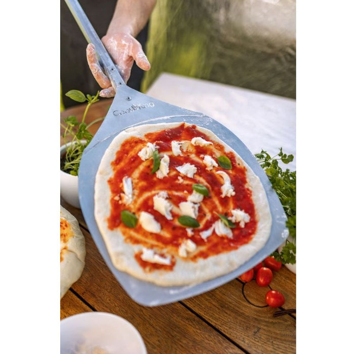 Närbild på pizzaspade med rå pizza toppad med tomatsås, mozzarella och basilika, redo att gräddas.
